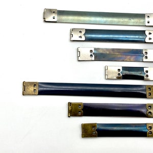 May include: Six metal strap buckles in various sizes and finishes. The buckles are silver and brass with a blue, black, and silver finish. The buckles are used for attaching straps to bags, purses, and other items.