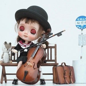 Könnte beinhalten: Eine Puppe in einem schwarz-weißen Matrosenkleid und einem schwarzen Hut spielt Cello, während sie auf einer Holzbank sitzt. Ein kleines Stofftier sitzt neben der Puppe auf der Bank. Ein blaues und weißes Bushaltestellenschild mit japanischem Text lautet "七国山分  福  前  バス停".