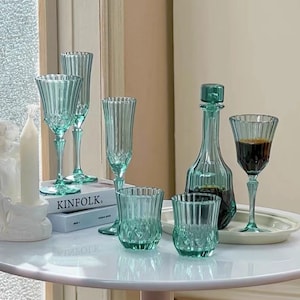 May include: A set of six green glass drinking glasses, including a decanter, on a white table. The glasses have a ribbed design and are arranged in a group. The decanter is tall and slender with a round base. The table is round and has a white surface.