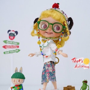 May include: A yellow-haired doll with green glasses and a white shirt is wearing a colorful beaded necklace. The doll is holding a sign that says "That's hilarious" and is standing next to a small rabbit doll and a green frog doll.