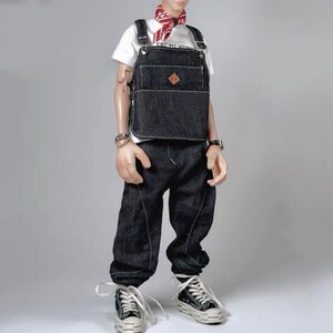 May include: A denim overall vest with a brown leather patch and a white t-shirt underneath. The model is wearing black jeans and white sneakers.