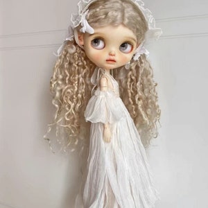 Può includere: Una bambola con lunghi capelli biondi ricci che indossa un vestito bianco e un berretto di pizzo bianco.