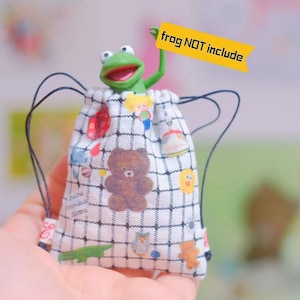 Puede incluir: Una bolsa de cordón blanco con un patrón de cuadrícula negra y personajes de dibujos animados coloridos impresos en ella. La bolsa está hecha de tela y tiene un cierre de cordón. La bolsa está siendo sostenida en una mano. "frog NOT include" está escrito en una pancarta amarilla.