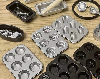 May include: A set of miniature baking pans in silver and black. The pans are in various shapes, including muffin tins, loaf pans, and cupcake pans. The pans are arranged on a wooden surface.