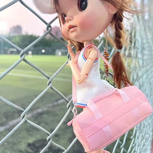 以下が含まれることがあります： 白いドレスに青と赤のアクセント、白い帽子、ビーズのストラップが付いたピンクのダッフルバッグを身に着けた人形。人形は茶色の髪を三つ編みにし、鎖のフェンスの近くに立っています。