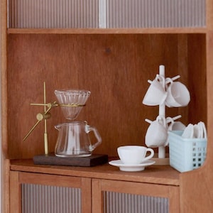 May include: A wooden cabinet with glass doors and shelves. The top shelf has a glass coffee dripper, a glass carafe, and a gold metal stand. The bottom shelf has a white ceramic cup and saucer, a white ceramic cup rack with white cups, and a light blue plastic basket.