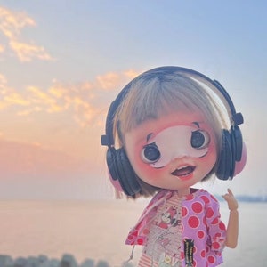 Può includere: Una bambola con una testa grande e occhi espressivi che indossa le cuffie e una giacca rosa a pois. La bambola è ambientata su uno sfondo di tramonto sull'acqua, con un sole rosso brillante visibile.