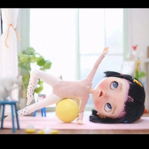 May include: A doll wearing a pink leotard and white tights with black polka dots is doing a side plank exercise with a yellow exercise ball. The doll has black hair with a pink headband that says "JA" on it. The doll is on a pink yoga mat.