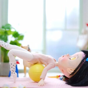 May include: A doll wearing white tights and a pink leotard is lying on a yellow exercise ball. The doll has black hair with a blue bow and is looking up. The doll is on a pink yoga mat.