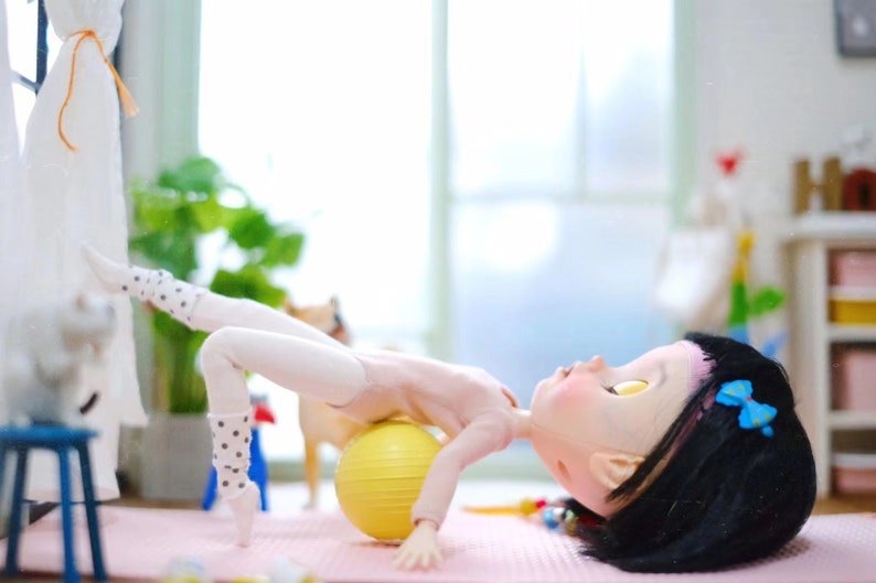 May include: A doll wearing white tights and a pink leotard is lying on a yellow exercise ball. The doll has black hair with a blue bow and is looking up. The doll is on a pink yoga mat.