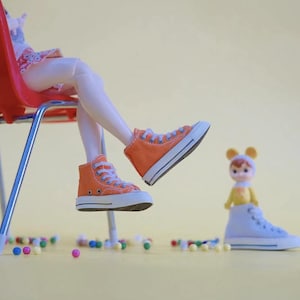 May include: A doll wearing a white dress and orange high-top sneakers sits on a red chair. The doll's legs are extended and one foot is pointing towards a smaller doll wearing a yellow shirt and white high-top sneakers.