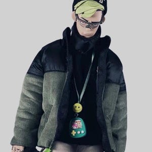 Pode incluir: Uma jaqueta puffer verde e preta com uma gola alta preta por baixo. A jaqueta tem um fecho de correr e um painel de veludo cotel&ecirc; verde na frente. A pessoa est&aacute; usando um amuleto de cara sorridente verde e amarelo em um cord&atilde;o ao redor do pesco&ccedil;o.