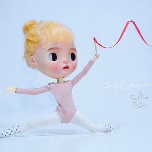 May include: A doll wearing a pink leotard and white tights with black polka dots is holding a red ribbon. The doll has blonde hair and is posed in a split. The background is a light blue. The text "Afternoon. Follow Me" is written in white on the right side of the image.