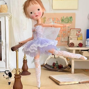 May include: A ballerina doll in a light purple tutu and white lace stockings, posed at a ballet barre. The doll has a delicate face and is wearing a white lace top. The ballet barre is made of wood and brass.