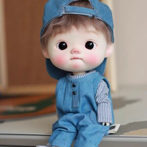 May include: A small, blue denim doll with a blue baseball cap, wearing a blue denim overall and a striped shirt. The doll has brown hair and large, dark eyes.