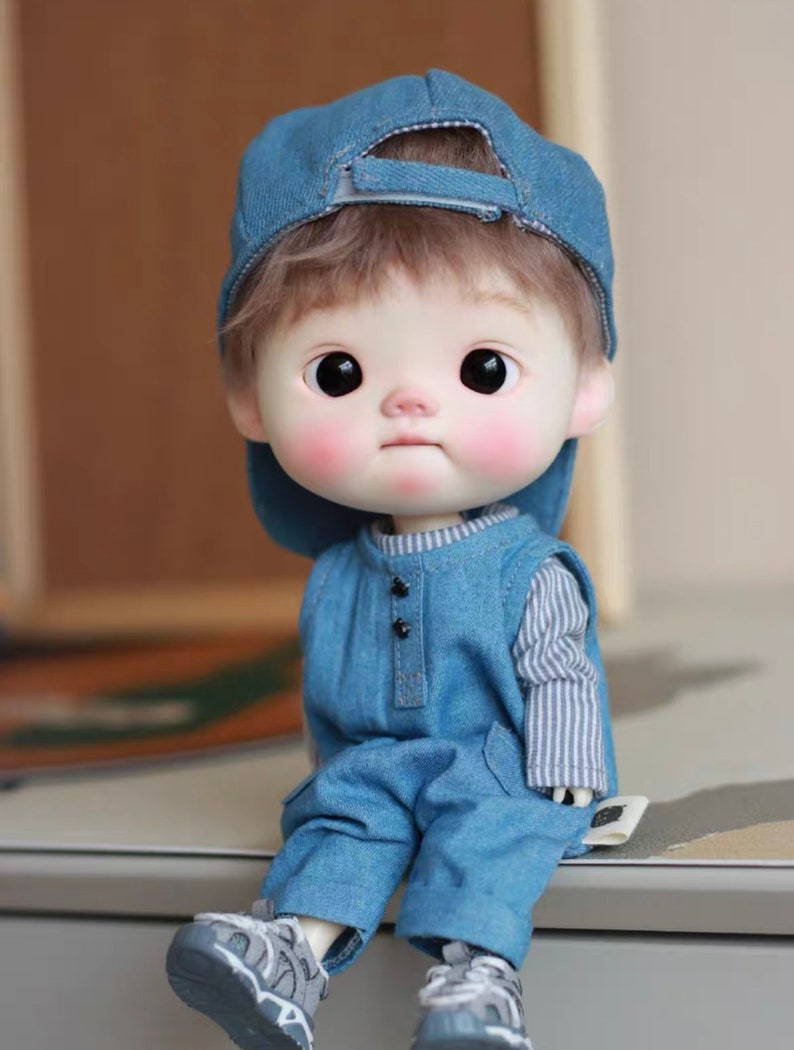 May include: A small, blue denim doll with a blue baseball cap, wearing a blue denim overall and a striped shirt. The doll has brown hair and large, dark eyes.