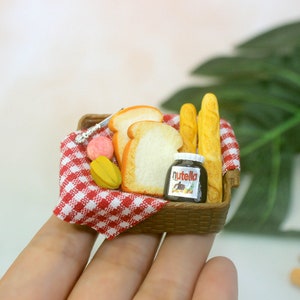 May include: A miniature picnic basket with a red and white checked cloth, containing sliced bread, baguettes, a jar of Nutella, and fruit. A small knife and pink and yellow fruit are also included. The basket is brown.