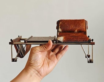 May include: A miniature chair and table set with a brown and white fur seat and backrest. The set has a wooden frame and a metal base. The table has a wooden surface. The set is held in a hand.