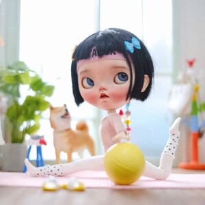 May include: A doll with black hair and a pink headband is wearing a white and black polka dot outfit and holding a yellow ball. The doll is sitting on a pink mat with a white background.