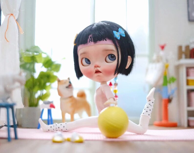 May include: A doll with black hair and a pink headband is wearing a white and black polka dot outfit and holding a yellow ball. The doll is sitting on a pink mat with a white background.