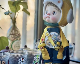 May include: A small doll wearing a yellow and green hat with a white brim and a yellow and blue jumpsuit with a cartoon character print. The doll is holding a yellow toy and is standing on a table with three clear glasses with painted designs.