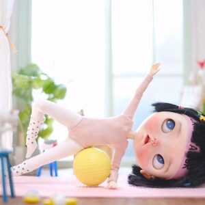 May include: A doll wearing a pink leotard and white tights with black polka dots is doing a side plank exercise on a yellow exercise ball. The doll has black hair and blue eyes and is wearing a pink headband with the text "A" on it.