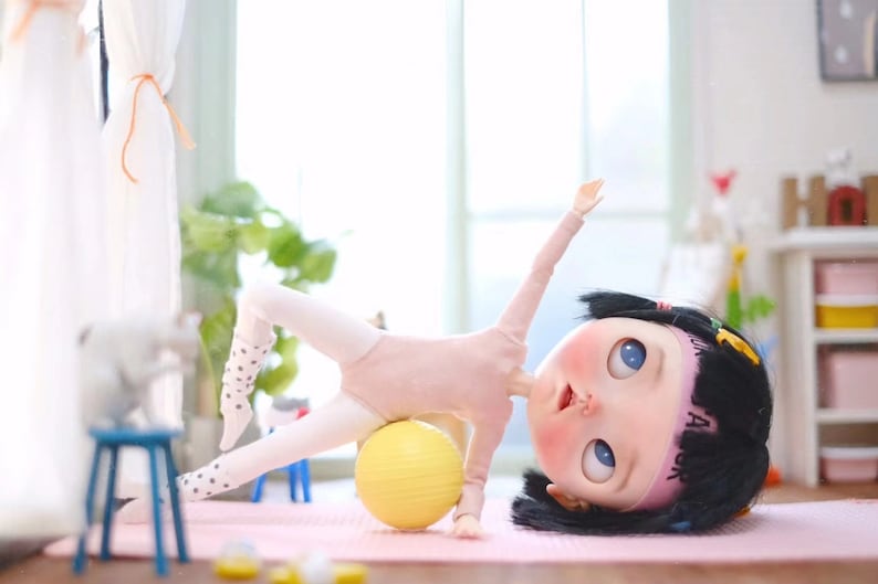 May include: A doll wearing a pink leotard and white tights with black polka dots is doing a side plank exercise on a yellow exercise ball. The doll has black hair and blue eyes and is wearing a pink headband with the text "A" on it.