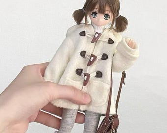 Könnte beinhalten: Eine Puppe in einem cremefarbenen Dufflecoat mit braunen Knebelverschlüssen, einem weißen Rollkragenpullover und grauen Leggings. Die Puppe hat braune Zöpfe und trägt eine kleine braune Umhängetasche. Weiße Socken und Stiefel vervollständigen das Outfit.