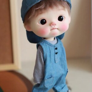 May include: A small doll wearing a blue denim overall with a striped shirt underneath. The doll is wearing a blue baseball cap and has brown hair. The doll is standing on a white surface.