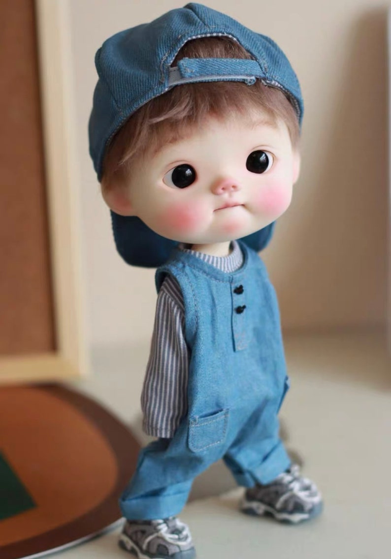 May include: A small doll wearing a blue denim overall with a striped shirt underneath. The doll is wearing a blue baseball cap and has brown hair. The doll is standing on a white surface.