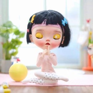 May include: A black-haired doll with yellow eyes is sitting in a yoga pose on a pink mat. The doll is wearing a pink long-sleeved shirt, white tights with black polka dots, and a pink headband with the word "JUICY" on it. A yellow exercise ball is in the background.