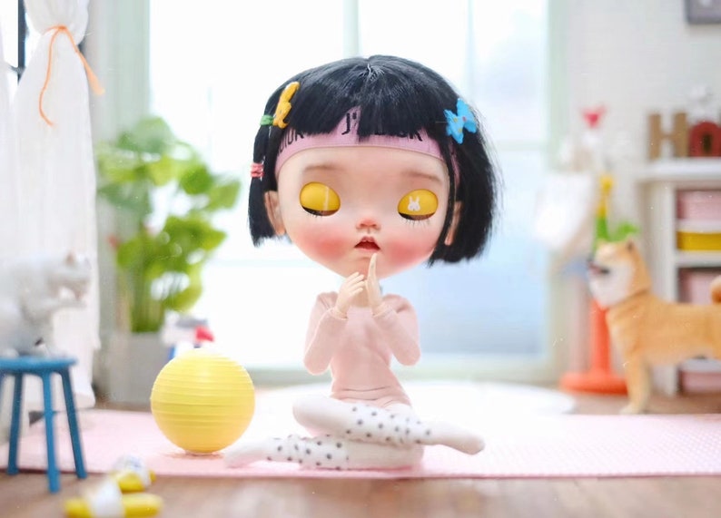 May include: A black-haired doll with yellow eyes is sitting in a yoga pose on a pink mat. The doll is wearing a pink long-sleeved shirt, white tights with black polka dots, and a pink headband with the word "JUICY" on it. A yellow exercise ball is in the background.