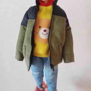 Pode incluir: Uma jaqueta de veludo cotel&ecirc; verde, preta e amarela com fecho de correr. A jaqueta tem um capuz amarelo com um gr&aacute;fico de urso castanho na frente. A jaqueta &eacute; usada sobre cal&ccedil;as de ganga azuis.