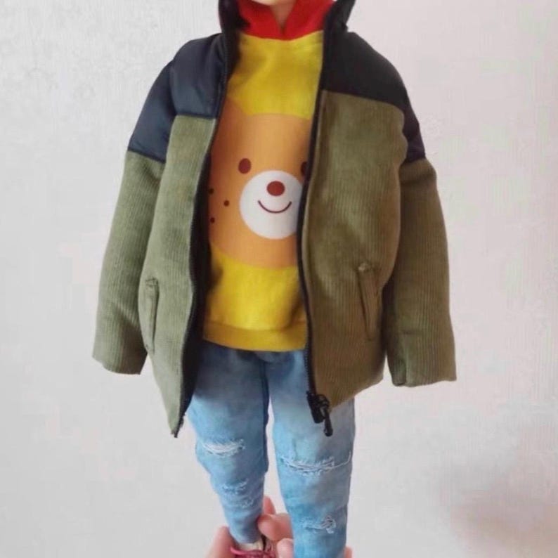 Pode incluir: Uma jaqueta de veludo cotel&ecirc; verde, preta e amarela com fecho de correr. A jaqueta tem um capuz amarelo com um gr&aacute;fico de urso castanho na frente. A jaqueta &eacute; usada sobre cal&ccedil;as de ganga azuis.