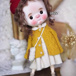 May include: A doll with curly brown hair wearing a yellow knitted cardigan, a white and yellow gingham dress, and a white crocheted bag. The doll is wearing a red beret and has a painted face with rosy cheeks.