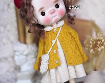 Könnte beinhalten: Eine Puppe mit lockigem braunen Haar, die einen gelben Strickcardigan, ein weiß-gelbes Gingham-Kleid und eine weiße gehäkelte Tasche trägt. Die Puppe trägt eine rote Baskenmütze und hat ein bemaltes Gesicht mit rosigen Wangen.