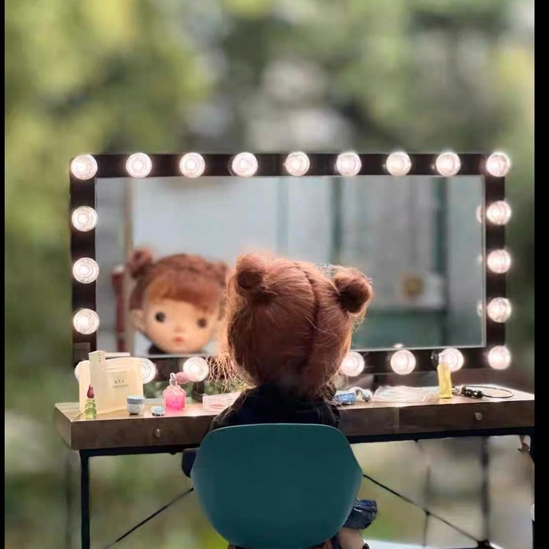May include: A doll with brown hair sits in front of a vanity mirror with lights. The mirror has a black frame and is surrounded by white lights. The vanity table is made of wood and has a light brown finish. There are several bottles and jars on the table.