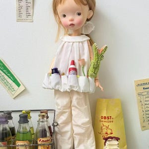 Può includere: Una bambola vestita con un top bianco con tasche e pantaloni color crema, che tiene in mano un giocattolo a forma di coccodrillo verde. Una borsa gialla con la scritta "OBSTu Gemüse" è dietro la bambola. Bottiglie di liquido in un rack di metallo.