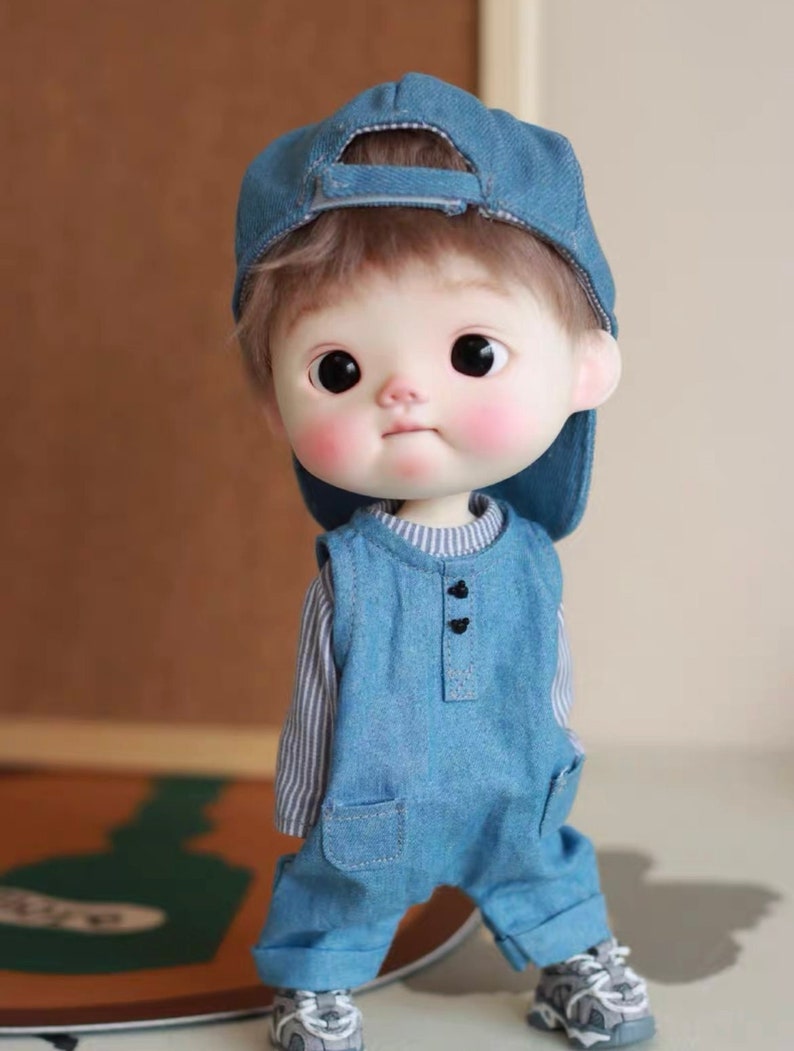May include: A small doll wearing a blue denim overall, a blue denim baseball cap, and grey sneakers. The doll has brown hair and a painted face with rosy cheeks.
