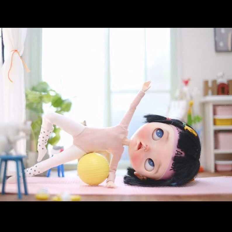 May include: A doll wearing a pink leotard and white tights with black polka dots is doing a side plank exercise with a yellow exercise ball. The doll has black hair with a pink headband that says "JA" on it. The doll is on a pink yoga mat.