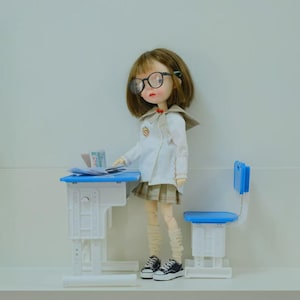 Pode incluir: Uma boneca vestida com uniforme escolar, usando óculos, ao lado de uma mesa e cadeira azuis e brancas. A boneca tem cabelo castanho curto e usa um casaco branco, uma saia xadrez e tênis preto e branco. Há papéis na mesa.