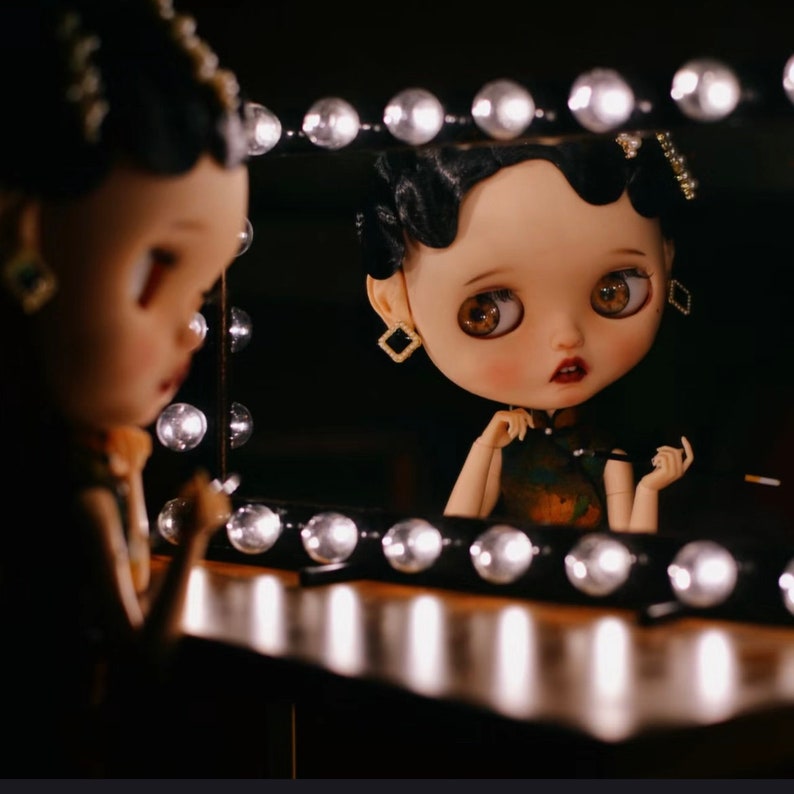 May include: A doll with black hair and a white dress is looking in a vanity mirror with light bulbs. The doll has a light pink face with brown eyes and a red mouth. The doll is wearing gold earrings.