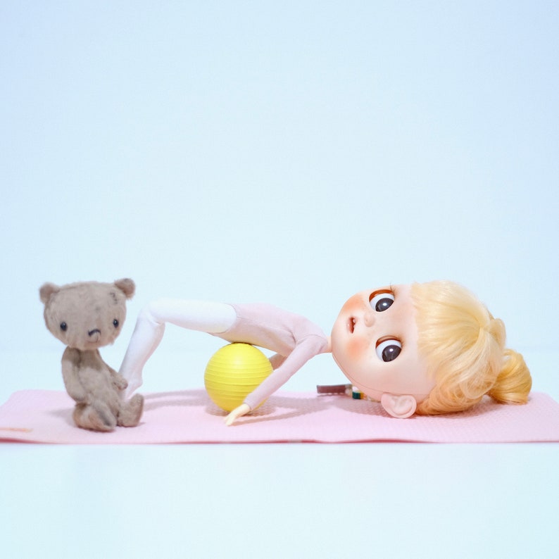 May include: A doll with blonde hair is lying on a pink yoga mat with a yellow exercise ball. A brown teddy bear is sitting on the mat to the left of the doll.