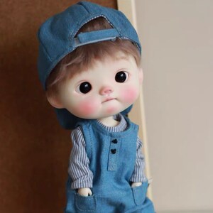 May include: A small doll with a blue denim cap, a striped long-sleeved shirt, and blue denim overalls. The doll has rosy cheeks and large brown eyes.