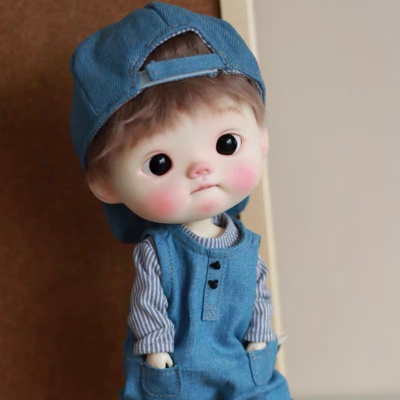 May include: A small doll with a blue denim cap, a striped long-sleeved shirt, and blue denim overalls. The doll has rosy cheeks and large brown eyes.