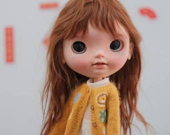 May include: A doll with long brown hair wearing a yellow cardigan with a white dress underneath. The cardigan has a lion and other animal designs on it.