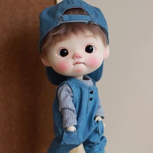 May include: A small doll with a round face, wearing a blue denim overall and a blue baseball cap. The doll has brown hair and large brown eyes.