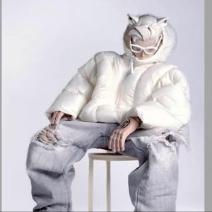 Puede incluir: Una chaqueta acolchada blanca con capucha y orejas de gato. La chaqueta tiene una cremallera y se lleva sobre unos vaqueros grises con detalles desgastados.