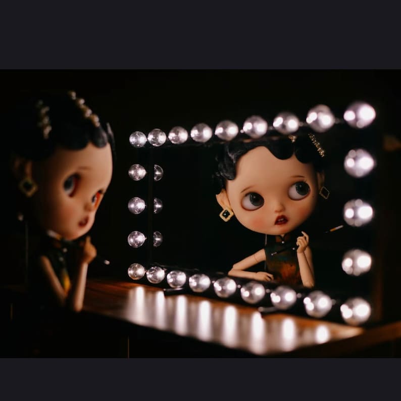 May include: A doll with black hair and a green and gold dress is looking into a vanity mirror with light bulbs around the frame.
