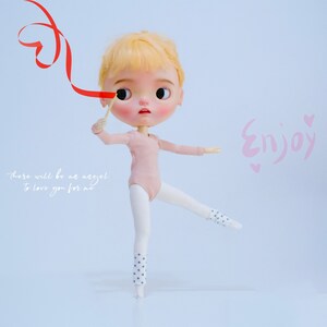 May include: A doll with blonde hair wearing a pink leotard and white tights with black polka dots is holding a red ribbon in the shape of a heart. The doll is standing on one leg with the other leg extended out to the side. The background is a light blue. The text "Enjoy" is written in pink on the right side of the image. The text "there will be an angel to love you for me" is written in white on the left side of the image.